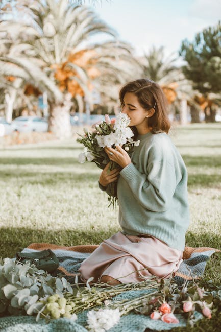 A young woman with shoulder-length brown hair and a gentle expression is sitting on a patterned blanket on the grass near Victoria Park. She is holding a fresh floral bouquet featuring white roses, pe
