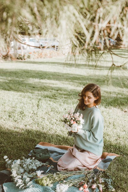 A young woman with shoulder-length brown hair, dressed in a light blue sweater and soft pink skirt, sits on a picnic blanket on a grassy area near Victoria Park. She is holding a bouquet of fresh pink