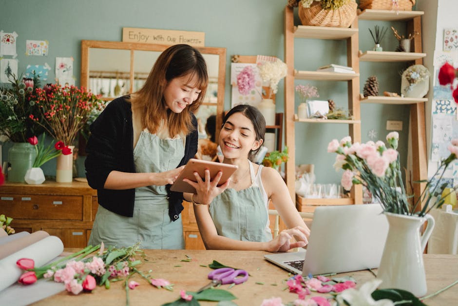 Two women in a flower shop, wearing light green aprons, are smiling and engaging with a tablet while surrounded by various fresh flowers. The woman on the left, with shoulder-length brown hair, holds 