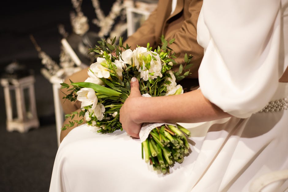 A close-up of a bride holding a bouquet of fresh white flowers, including roses and lisianthus, accented with green foliage. The bouquet features a natural, rounded arrangement wrapped with white lace