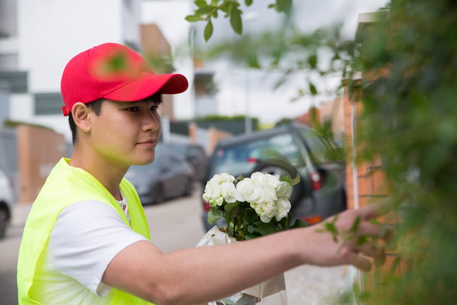 A young male florist with light skin, wearing a red cap and a bright yellow high-visibility vest over a white t-shirt, is holding a floral arrangement featuring white hydrangeas and green foliage. He 